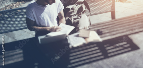 Stylish man wearing white tshirt sitting city park and reading book. Studying at the University, working new project. Books, laptop, backpack bench. Horizontal, blurred