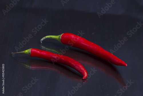 Two red hot chili peppers are on a mirror black wooden table