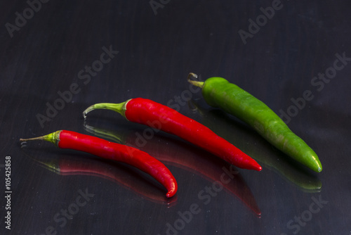 Two red and one green hot chili peppers are on a mirror black wooden table