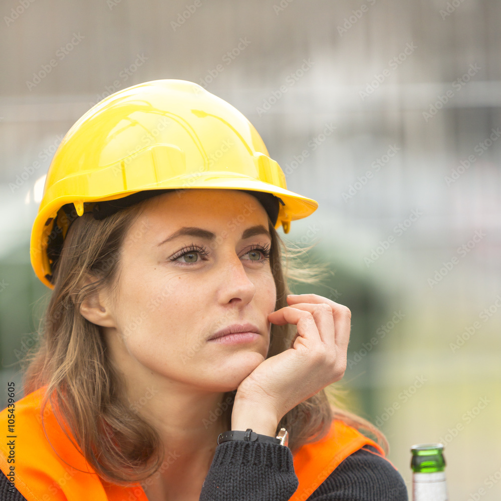 Pouting female engineer with safety jacket and helmet with beer during ...