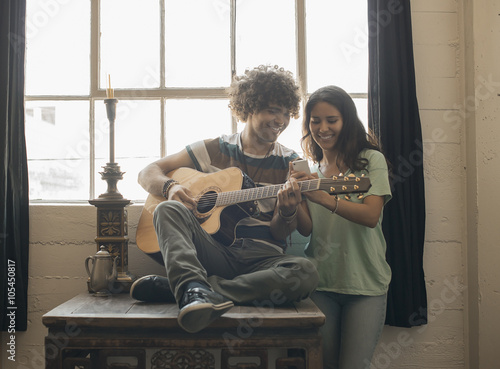 Loft living, A young man playing guitar and a woman beside him taking photographs with a smart phone,  photo