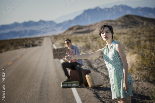 A young couple, man and woman, on a tarmac road in the desert hitchhiking, with a sign saying Vegas or Bust,