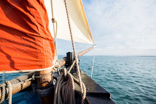 rope wound on a wooden cleat fixed on the hull of a rigging vintage sailing boat with a beige jib and an ocher sail filled by the wind at the blur background during a sunny sea trip in brittany photo