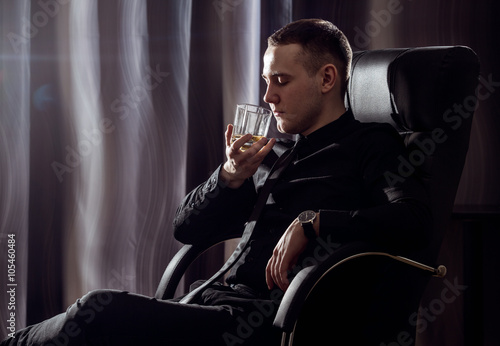 Bring down stress. Young handsome stressed businessman in formal wear taking off his necktie holding a glass of whiskey in his hand. Isolated on black background. photo