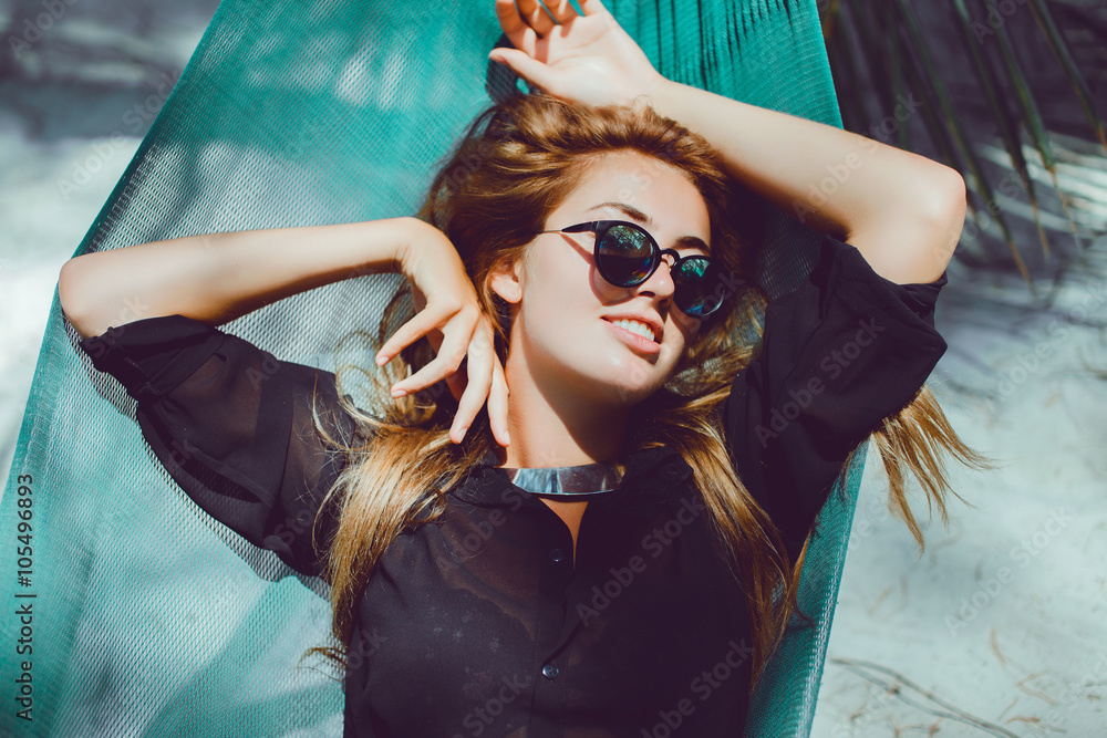 Obraz premium young beautiful girl in sunglasses denim shorts and shirt posing while lying on a hammock, beautiful woman, brunette resting on vacation at the beach, relax, outdoor portrait