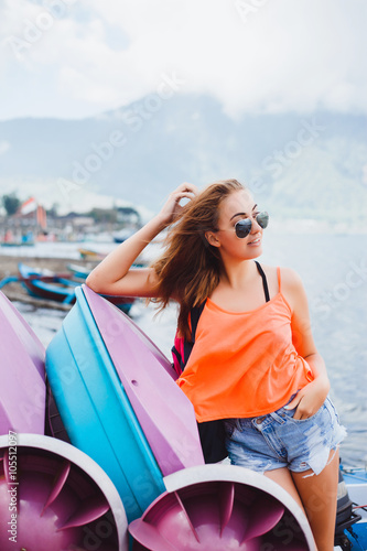 haired young girl in sunglasses and a red t-shirt denim shorts travels with a travel backpack for the world, posing next to a mountain lake, around the boats and boats