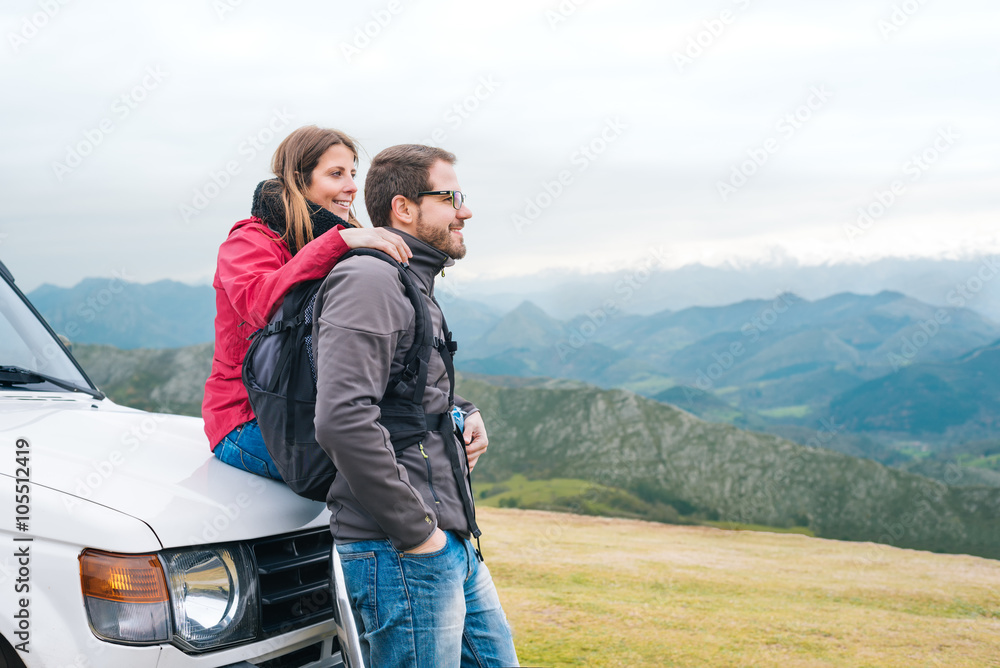 Young couple in love leaning on all terrain car, looking to the horizon, hiking outdoor on vacation, enjoying relaxing rural holidays.