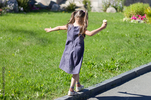 Little cute girl walking on the curb with ice cream in hand and intently trying to keep her balance, with her back