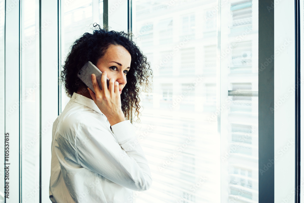 Young attractive businesswoman talking on mobile phone while standing near the window in the office center Young attractive businesswoman talking on mobile phone while standing near the window in the office center