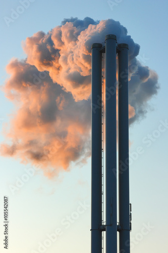 Emission into the environment of steam coming out of factory chimneys. Dense cloud of smoke in the red light of the sunlight.