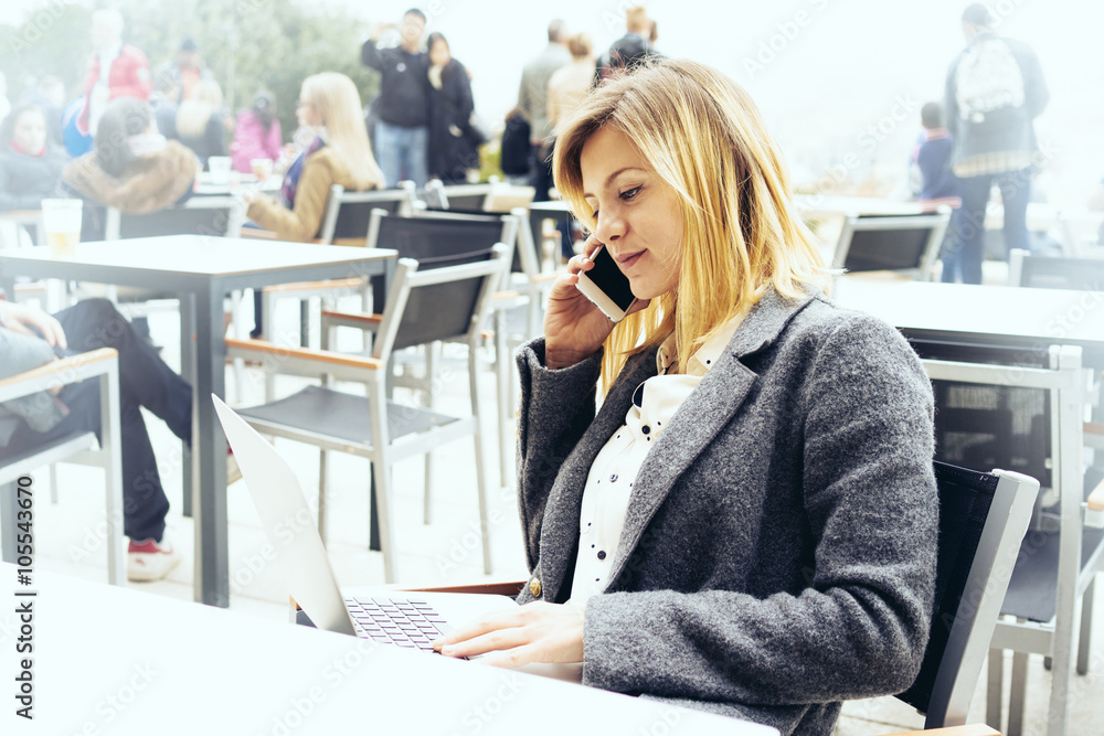 Portrait of a beautiful young businesswoman who is holding portable laptop computer while sitting in a coffee shop outdoors and talking on the smart-phone
