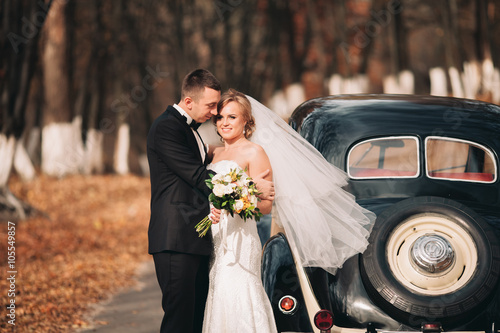Stylish wedding couple, bride, groom kissing and hugging near retro car in autumn
