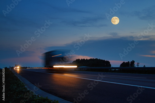 Trucks with glowing lights arriving on the motorway from afar in the landscape with a full moon at dusk. Motion blur speeding trucks. Dark blue summer sky with clouds and alpenglow.