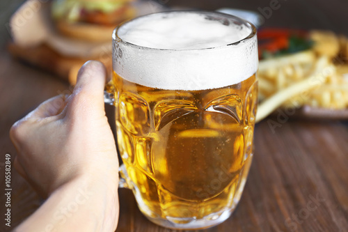 Hand holding glass mug of light beer with snacks on dark wooden table, close up