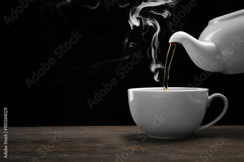 Pouring hot tea from a kettle into a cup on black background, close up