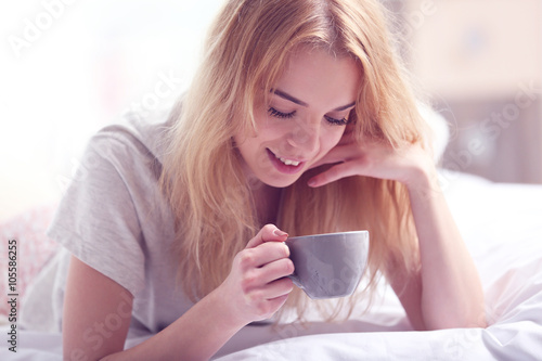 Attractive young blonde girl in a pajama holding a cup while laying in bed