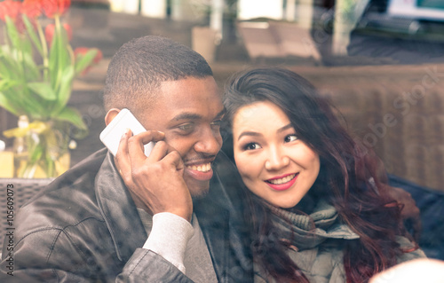 Multiracial couple on date view through cafe window - Afro american man using phone and asian girlfriend facial expression in love - Cheerful mixed race friends - Closeup from glass focus on male 