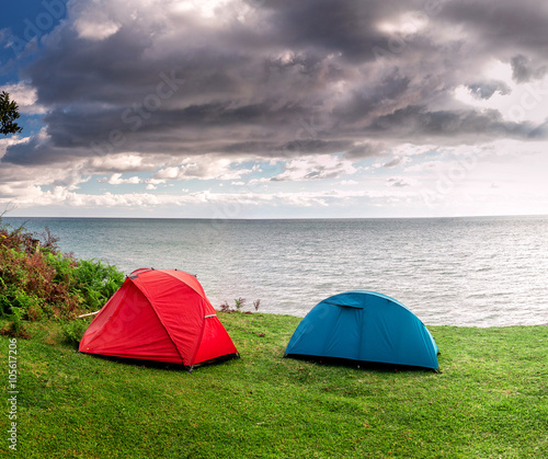 Red and blue tents are set on the grass by the sea
