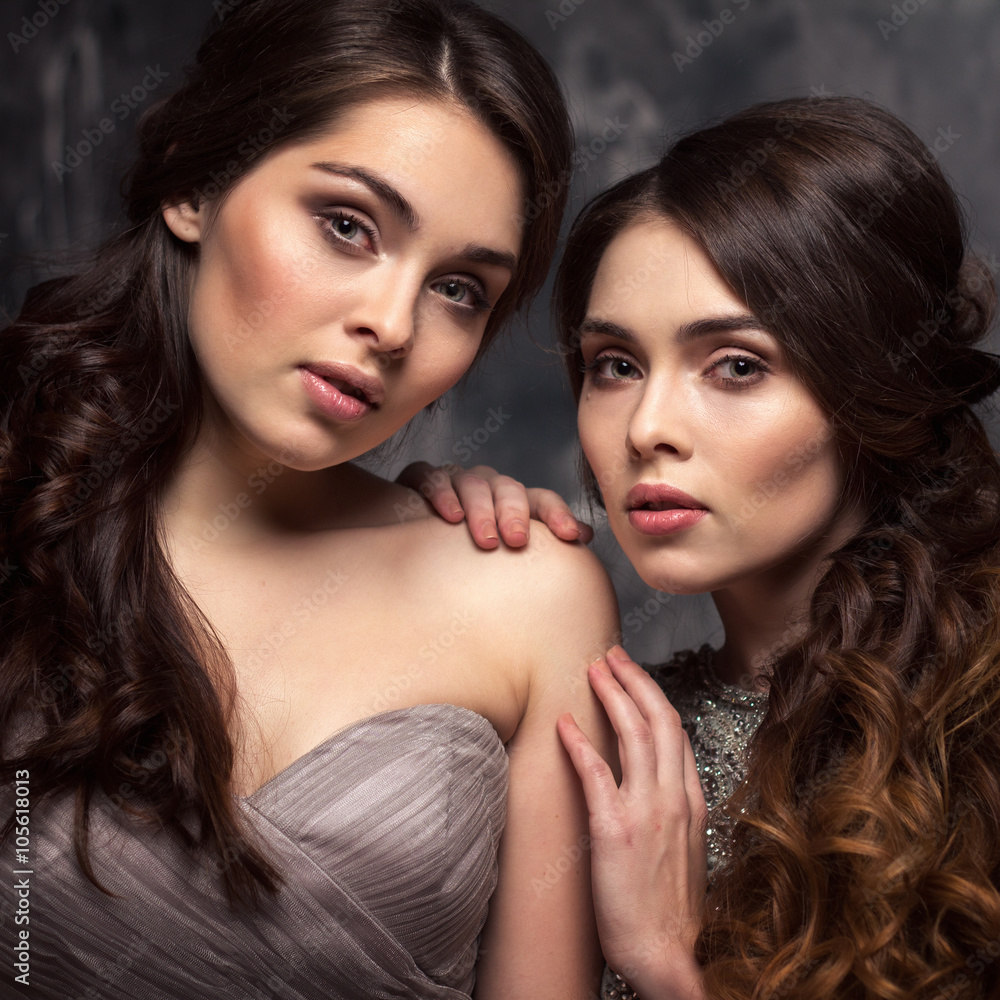 Fashion beauty portrait of gorgeous young twins women with long curly hair in luxury evening dresses