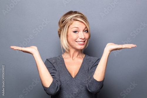 comparison concept - happy young blond woman displaying something on both flat hands for similar choice of product, gray background studio. photo