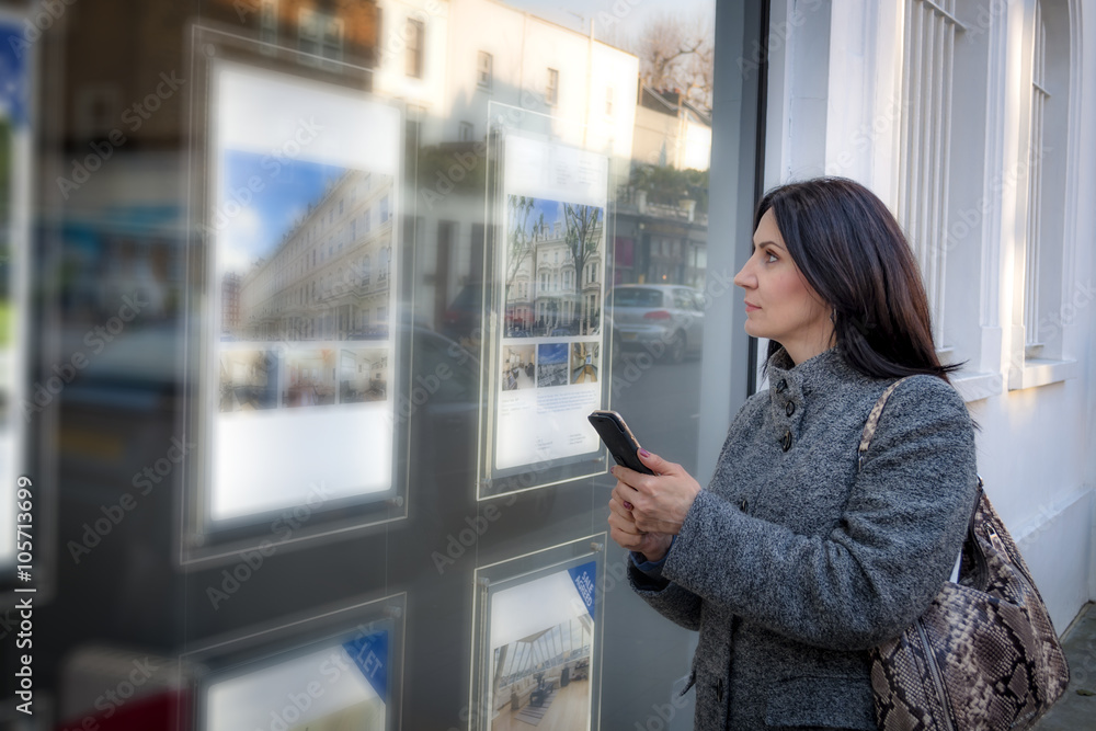 Mature woman wearing a grey coat and a purse is looking at the ads in the window of a real estate agency for a new place to live, and takes notes on her phone