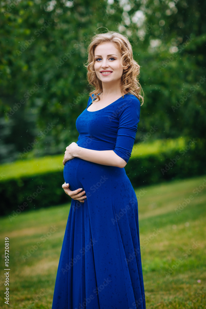 Adorable smiling young pregnant woman in blue dress with long blond curly hair holding her belly in summer park on rainy day. Pregnancy and femininity concept. Water drops on blue dress Adorable smiling young pregnant woman in blue dress with long blond curly hair holding her belly in summer park on rainy day. Pregnancy and femininity concept. Water drops on blue dress