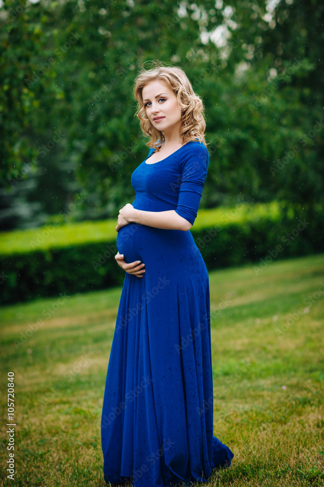 Pretty young pregnant woman in blue dress with long blond curly hair holding her belly and looking at camera in summer park on rainy day. Pregnancy and femininity concept. Water drops on blue dress Pretty young pregnant woman in blue dress with long blond curly hair holding her belly and looking at camera in summer park on rainy day. Pregnancy and femininity concept. Water drops on blue dress