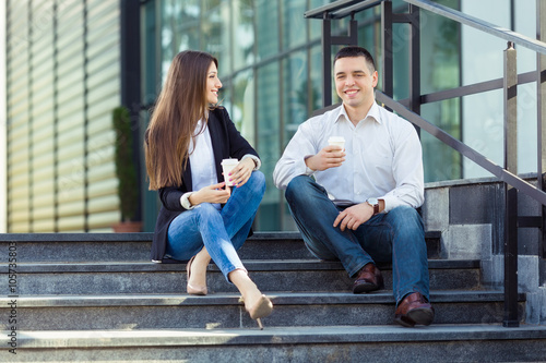 Young businesswoman and businessman sitting on the steps of the office building  drinking coffee and talking. Young man is looking at camera and young woman is looking at him.