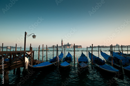 Pictorial view of blue covered gondolas and bridge with a streetlight in lagoon of Venice, Italy. Evening shot in soft natural colors with island and tower on background.