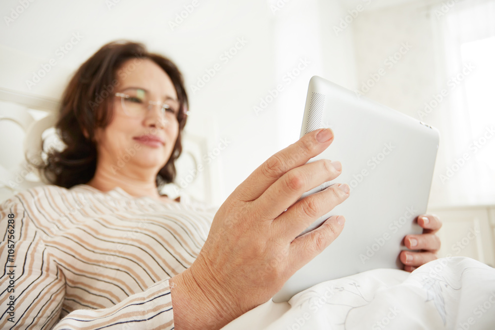 Attentive and cheerful mature woman using a tablet pc lying on her bed in a bedroom.