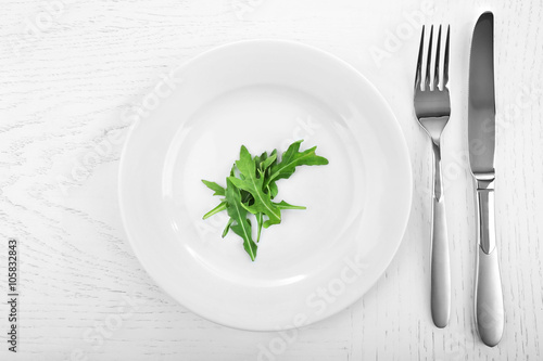 Arugula leaves in plate with fork and knife on wooden table, top view