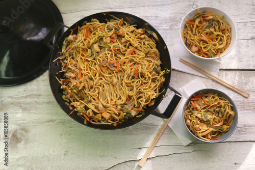 Noodles with chicken and various vegetables, prepared and served in wok. Two smaller bowls with chopsticks on the side. Served on white rustic table. Top view, selective focus. 
