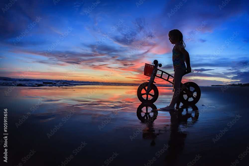 Fototapeta premium Black silhouette of little child - run bike rider stand on wet sand beach, look at sea surf and colourful sunset sky. Family travel lifestyle background, outdoor fun activity on Bali summer vacation.