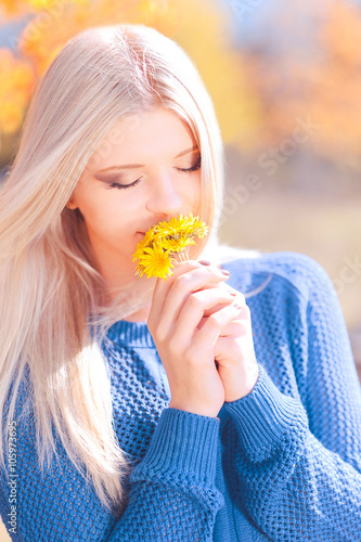 Beautiful blonde girl smelling dandelions outdoors over nature background. Eyes closed. Spring season. Sunny day. 20s. 
