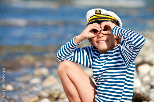 Boy in striped vest and a cap sailor holding hands around the eyes like a pair of binoculars at the sea shore