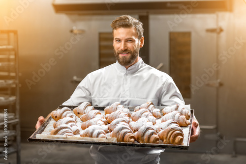 Slika na platnu Handsome baker in uniform holding tray full of freshly baked croisants at the ma
