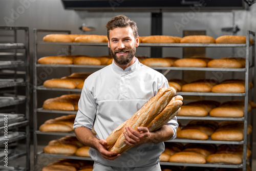 Leinwand Poster Handsome baker in uniform holding baguettes with bread shelves on the background