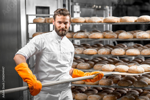Handsome baker in uniform with orange working gloves putting with shovel from the oven bread loafs on the shelves at the manufacturing