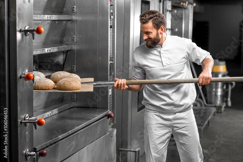 Handsome baker in uniform taking out with shovel freshly baked buckweat bread from the oven at the manufacturing photo