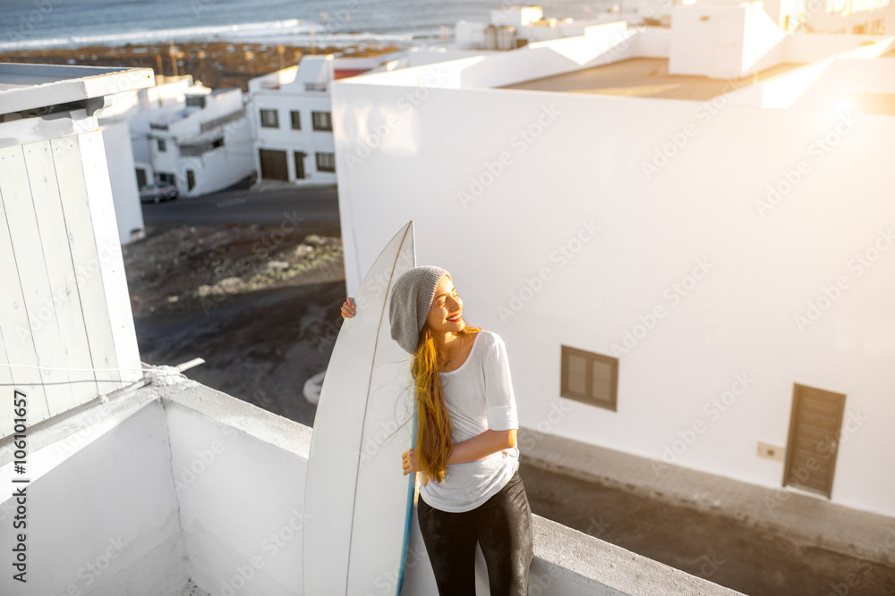 Young smiling woman in white t-shirt and hat sitting with surfboard on the roof top on the white city background. Enjoying morning sunlight and preparing for surfing Young smiling woman in white t-shirt and hat sitting with surfboard on the roof top on the white city background. Enjoying morning sunlight and preparing for surfing