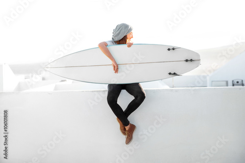 Photography Young smiling woman in white t-shirt and hat sitting with surfboard on the roof top on the white city background