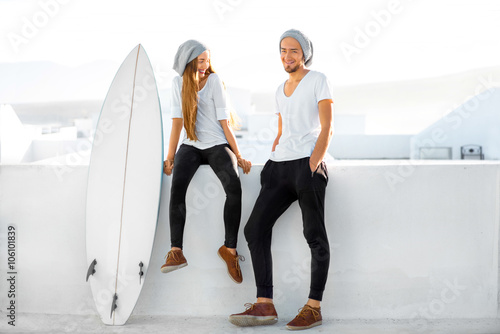 Young couple dressed alike in white t-shirts and hats enjoying summer vacations with surfboard on the roof top in the morning