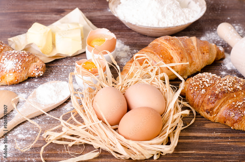 Ingredients for baking croissants - paper, flour, wooden spoon, rolling pin, eggs, egg yolks, butter served on a rustic wooden tray table. Ingredients for baking croissants - paper, flour, wooden spoon, rolling pin, eggs, egg yolks, butter served on a rustic wooden tray table.