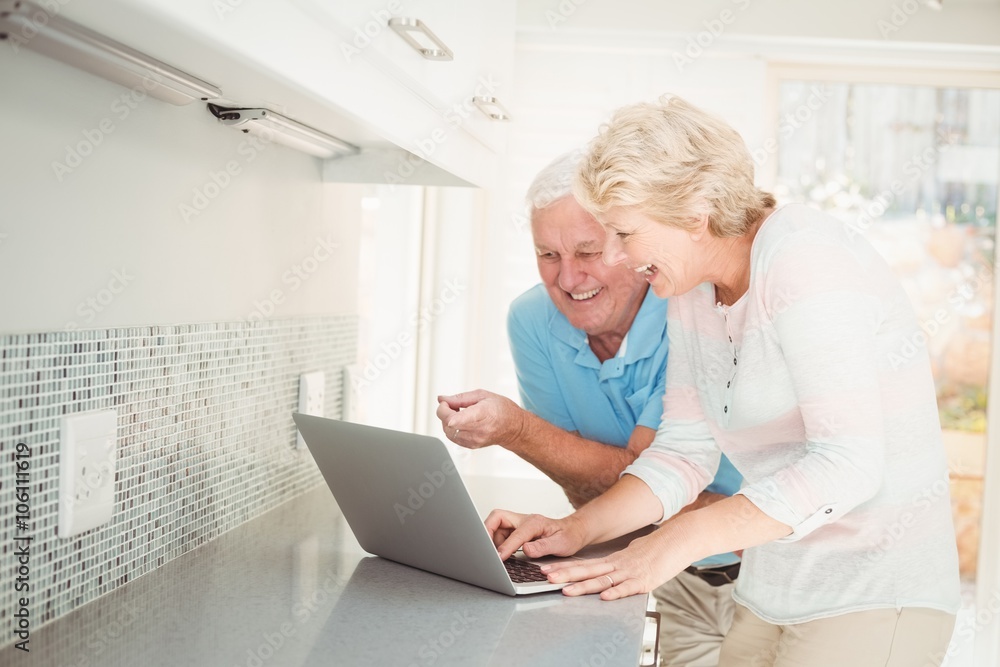 Senior couple laughing while using laptop in kitchen Senior couple laughing while using laptop in kitchen