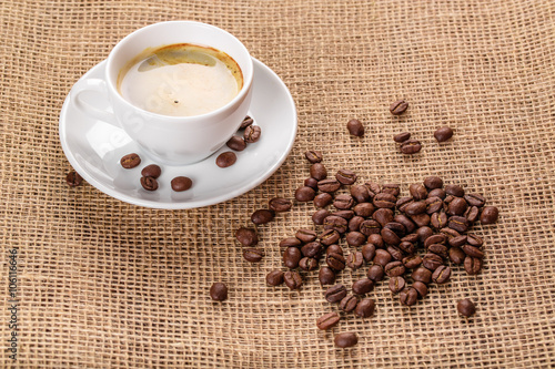 Beautiful white coffee cup and saucer and coffee beans on the background of burlap. Dark background.