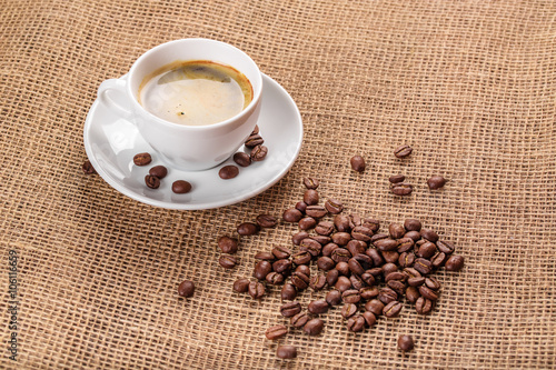 Beautiful white coffee cup and saucer and coffee beans on the background of burlap. Dark background.