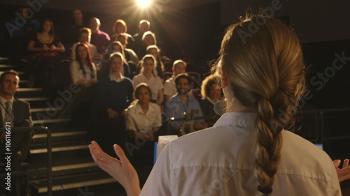 Over the shoulder view of a teenage girl giving a presentation to a group of students and teachers in her school.