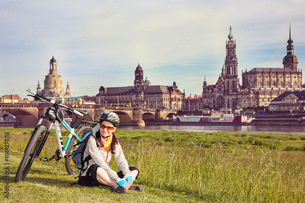 Obraz premium Adult attractive female cyclist sitting on the grass and resting. The happy woman sits at the old city background and looks at the camera. So-called The Balcony of Europe, Dresden, Germany