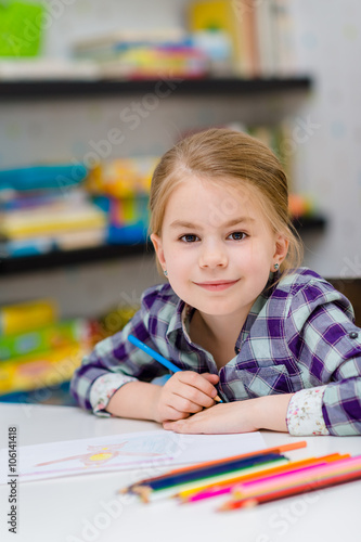 Lovely smiling little girl with blond hair sitting at white table with multicolored pencils and looking at camera
