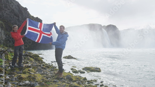 Happy young couple holding Iceland flag against Godafoss waterfall. Portrait of cheerful tourist couple enjoying vacation at beautiful waterfall. Male and female are visiting famous attractions. photo